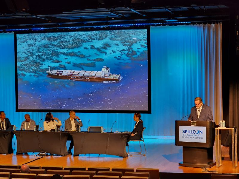 Man talking at podium right of screen. Projector with boat on screen sits behind people sitting at desk on a stage in an auditorium at Spillcon 2023